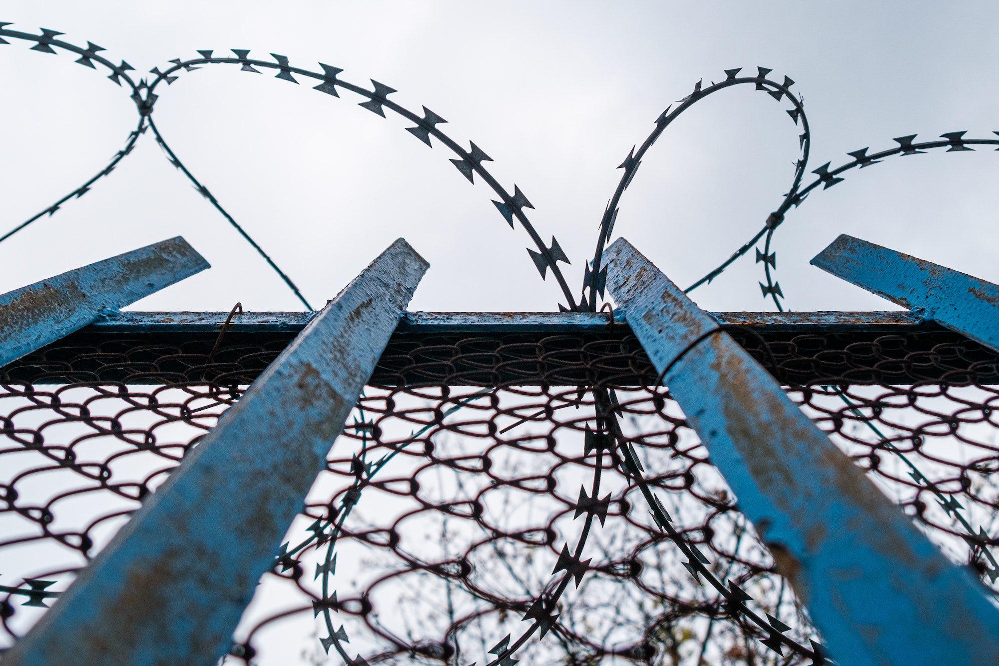 A blue fence with barbed wire near prison or mental hospital on the background of grey sky